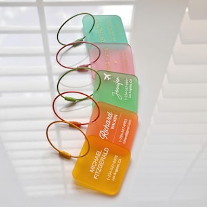 May include: A stack of colorful luggage tags in shades of green, pink, orange, and yellow. Each tag has a matching cord and features a name and contact information in white text. The tags are rectangular with rounded corners.