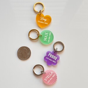 Op de afbeelding: Vier acryl huisdierlabels met metalen ringen in hart-, cirkel-, bloem- en geschulpte vormen. De kleuren zijn oranje, groen, paars en roze. Elk label is bedrukt met een witte naam en het telefoonnummer 123-456-7890. Een Amerikaanse munt dient als schaal.