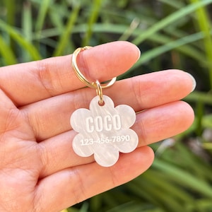 May include: A flower-shaped pet ID tag made of white pearlescent acrylic with a gold-tone metal split ring. The tag features the name COCO engraved in white capital letters above the phone number 123-456-7890 in a clean sans-serif font.