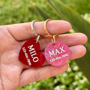 Puede incluir: Dos etiquetas de identificaci&oacute;n para mascotas. Una es en forma de coraz&oacute;n, roja, con el nombre "MILO" y un n&uacute;mero de tel&eacute;fono. La otra es una etiqueta rosa, circular, con el nombre "MAX" y un n&uacute;mero de tel&eacute;fono. Ambas tienen llaveros de metal.