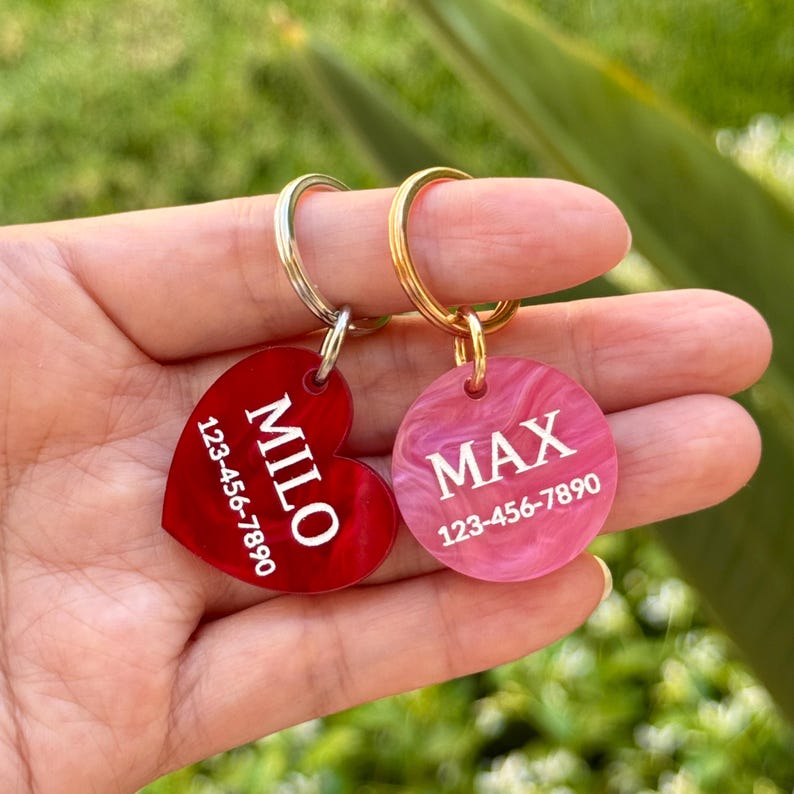 Puede incluir: Dos etiquetas de identificaci&oacute;n para mascotas. Una es en forma de coraz&oacute;n, roja, con el nombre "MILO" y un n&uacute;mero de tel&eacute;fono. La otra es una etiqueta rosa, circular, con el nombre "MAX" y un n&uacute;mero de tel&eacute;fono. Ambas tienen llaveros de metal.