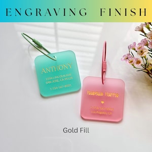 May include: Two square luggage tags, one teal and one pink, with gold lettering. The teal tag reads "ANTHONY" with an address and phone number. The pink tag reads "Isabela Harris" with an address and phone number. The text "ENGRAVING FINISH" is at the top.