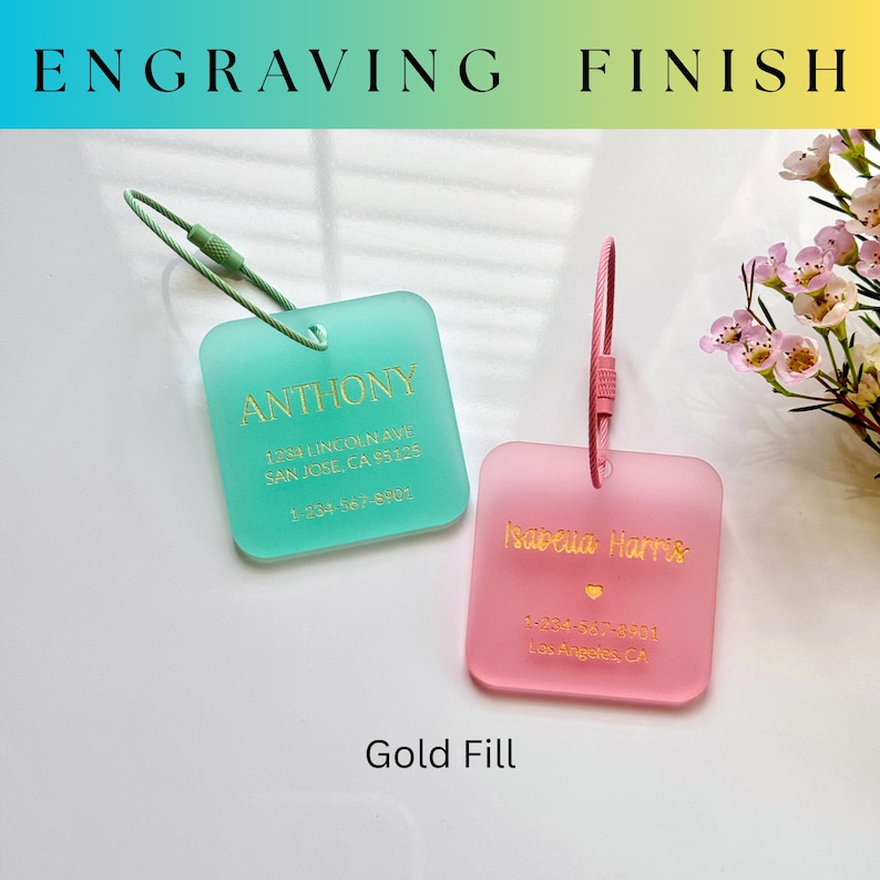 May include: Two square luggage tags, one teal and one pink, with gold lettering. The teal tag reads "ANTHONY" with an address and phone number. The pink tag reads "Isabela Harris" with an address and phone number. The text "ENGRAVING FINISH" is at the top.