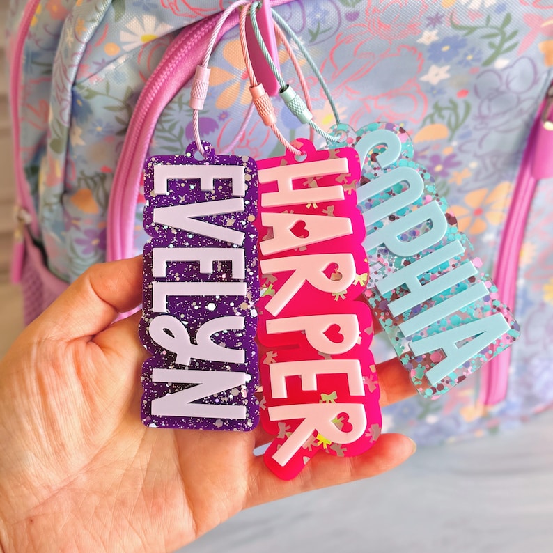 May include: Three personalized name tags in purple, pink, and blue with glitter accents. The tags read "EVELYN", "HARPER", and "SOPHIA" in white, with heart and butterfly details. They are attached to a floral backpack.