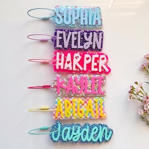 May include: A collection of personalized name tags in various colors and patterns. Each tag features a different name: Sophia, Evelyn, Harper, Kaylee, Abigail, and Jayden. The tags are made of clear acrylic with glitter and decorative accents.