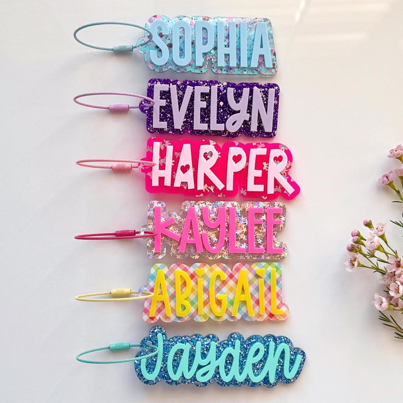 May include: A collection of personalized name tags in various colors and patterns. Each tag features a different name: Sophia, Evelyn, Harper, Kaylee, Abigail, and Jayden. The tags are made of clear acrylic with glitter and decorative accents.