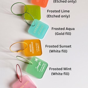 May include: A collection of frosted luggage tags in various colors, including orange, lime, aqua, sunset, mint, and pink. Each tag features etched or filled text with names and contact information. The tags are square with rounded corners and attached to metal loops.