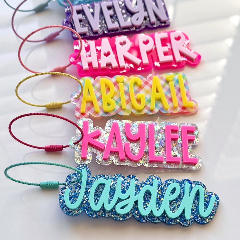 May include: A collection of colorful name tags with various names, including "Evelyn," "Harper," "Abigail," "Kaylee," and "Jayden." Each tag features a unique design, with glitter, hearts, and different color schemes. They are attached to metal loops.