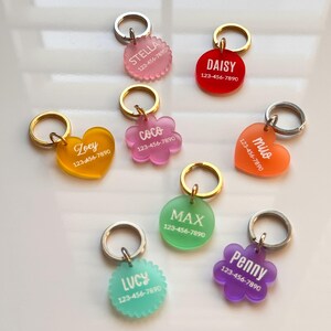 Op de afbeelding: Een verzameling van acht kleurrijke acryl naamplaatjes voor huisdieren in diverse vormen zoals cirkels, harten en bloemen. Elk plaatje bevat een witte naam en het telefoonnummer 123-456-7890, bevestigd aan een gouden of zilveren metalen ring.