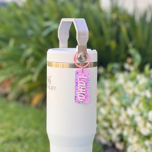 K&ouml;nnte beinhalten: Ein wei&szlig;er Isolierbecher mit grauem Griff und einem rosa Schl&uuml;sselanh&auml;nger. Der Schl&uuml;sselanh&auml;nger tr&auml;gt den Namen "Layla" in wei&szlig;er Schrift auf einem rosa-lila Glitzerhintergrund. Der Becher hat ein silbernes Logo.