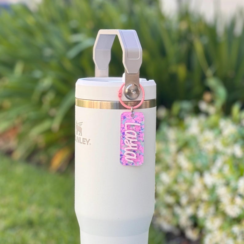 K&ouml;nnte beinhalten: Ein wei&szlig;er Isolierbecher mit grauem Griff und einem rosa Schl&uuml;sselanh&auml;nger. Der Schl&uuml;sselanh&auml;nger tr&auml;gt den Namen "Layla" in wei&szlig;er Schrift auf einem rosa-lila Glitzerhintergrund. Der Becher hat ein silbernes Logo.