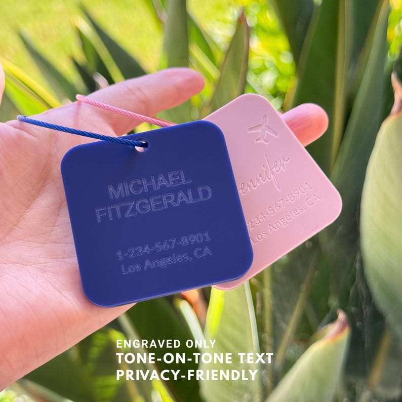 May include: Two personalized luggage tags. One is navy blue with the name "MICHAEL FITZGERALD" and a phone number. The other is pink with the name "Jennifer" and a phone number. The tags are engraved with tone-on-tone text and are privacy-friendly.