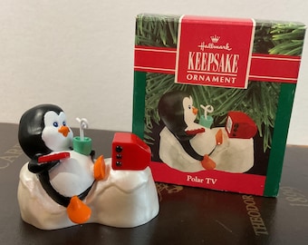 May include: A Hallmark Keepsake ornament featuring a penguin relaxing on a snow-covered surface, watching a red television. The penguin is black and white with orange feet and a green drink. The box reads "Polar TV".