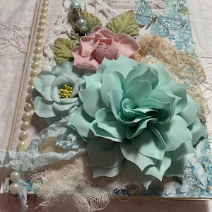 May include: A decorative journal cover featuring a large mint green paper flower, a smaller light blue flower with yellow centers, and a soft pink paper rose. The cover is embellished with white lace, cream-colored pearls, and a glittery blue butterfly.
