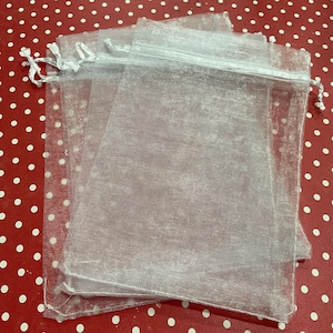May include: A stack of four sheer white organza drawstring bags. The bags are made of a translucent, shimmering fabric and are tied with a white satin ribbon. The bags are on a red surface with white polka dots.