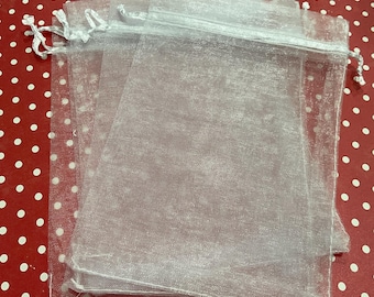 May include: A stack of four sheer white organza drawstring bags. The bags are made of a translucent, shimmering fabric and are tied with a white satin ribbon. The bags are on a red surface with white polka dots.