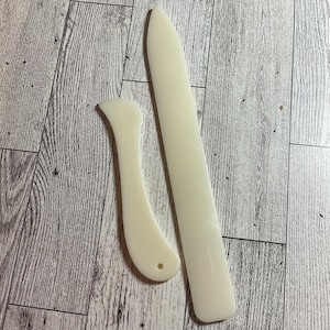 May include: Two ivory-colored bone folders on a wooden surface. One is a long, straight tool with a pointed end, and the other is curved with a rounded end and a small hole. These tools are used for paper crafting and bookbinding.