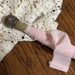 May include: A vintage-style bottle stopper with a wooden handle and metal accents. A pink fabric ribbon is wrapped around the handle. The stopper rests on a white crocheted doily, with a wooden surface in the background.