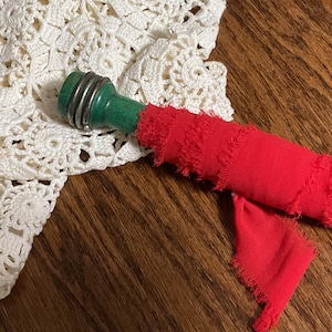 May include: A green and silver bottle stopper with a red fabric accent. The stopper has a wooden handle and a silver metal band. The red fabric is frayed at the edges. A white crocheted doily is in the background.