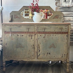 May include: A distressed, antique-style buffet with a mirror, painted in shades of green and brown. A white pitcher with red flowers sits on top. The buffet has two drawers and two cabinet doors.