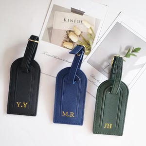 Könnte beinhalten: Drei Leder-Gepäckanhänger in Schwarz, Marineblau und Grün. Jeder Anhänger hat eine goldfarbene Schnalle und goldfarbene Buchstaben mit den Initialen: Y.Y, M.R und JH. Die Anhänger sind auf einer weißen Oberfläche mit dekorativem Papier und floralen Elementen angeordnet.