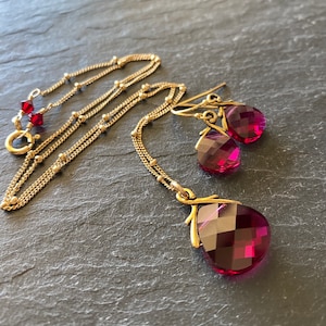 May include: A gold-colored necklace and matching earrings set. The necklace features a faceted, teardrop-shaped, magenta pendant. The earrings have matching magenta teardrop-shaped gems. The set is displayed on a dark gray surface.