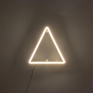 Op de afbeelding: Een driehoekige neonlamp die een warm wit licht uitstraalt. De lamp heeft de vorm van een gelijkzijdige driehoek met afgeronde hoeken en is aan een muur bevestigd. Een dunne zwarte stroomkabel komt uit de onderkant van het frame.