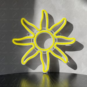 May include: A yellow neon sun-shaped light fixture. The light has a white frame and casts a shadow on a gray wall. The fixture is on a dark surface. The text "NAT NEST" is visible.