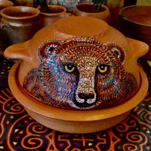 May include: A terracotta pot with a painted bear face on the lid. The pot is brown with a light brown rim and a handle on each side. The bear's face is brown with white fur and yellow eyes.