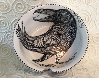 May include: A white ceramic bowl with a black and white illustration of a crow holding a white egg in its beak. The bowl has a black dotted rim.