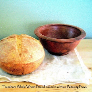 May include: A loaf of whole wheat bread baked in a brown clay bowl. The bread is light brown and has a slightly rough texture. The bowl is sitting on a white cloth.