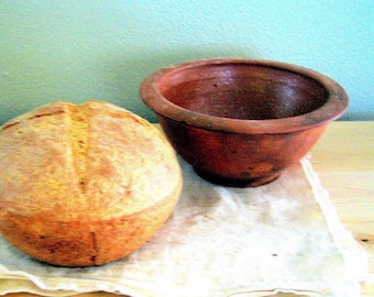 Puede incluir: Una hogaza de pan integral horneada en un bol de barro marrón. El pan es de color marrón claro y tiene una textura ligeramente rugosa. El bol está sobre un paño blanco.