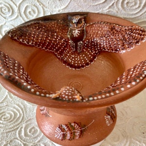 May include: A terracotta bowl with a brown and white owl design. The bowl has a wide rim and a narrow base. The owl is painted in detail with white dots and brown feathers.