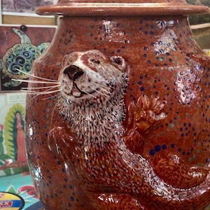 May include: A glossy, reddish-brown ceramic jar with a matching lid, featuring a raised, three-dimensional sculpture of an otter on the front. The otter is painted with white and brown details and has long, thin, white whiskers extending outward.
