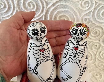 Op de afbeelding: Twee witte keramische zout- en pepervaatjes in de vorm van afgeronde figuren, handbeschilderd met een zwart skeletontwerp, een klein rood hartje op de borst en gedetailleerde gelaatstrekken. Eén figuur heeft een bloemenkrans in oranje en groen.