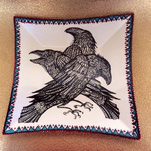 May include: A white square ceramic dish with a black and white illustration of two ravens. The dish has a decorative border with red, blue, and white triangles.