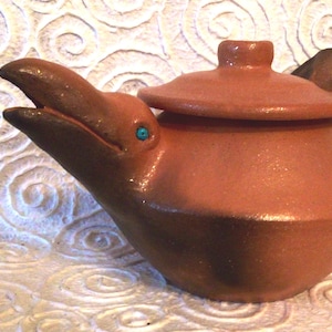 May include: A terracotta teapot shaped like a bird with a turquoise blue eye. The teapot has a lid and a spout. The teapot is sitting on a white surface.