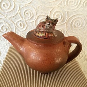 May include: A terracotta teapot with a fox figurine on the lid. The teapot has a speckled brown glaze and a gold-colored accent on the lid.