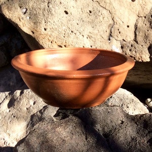 May include: A terracotta-colored ceramic bowl with a slightly rounded rim. The bowl is sitting on a pile of rocks.