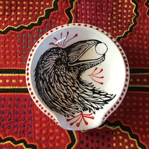 May include: A white ceramic spoon rest with a black and white illustration of a crow with its beak open. The crow is surrounded by red and white floral designs.