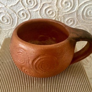 May include: A terracotta mug with a brown handle and a swirling pattern. The mug is made of clay and has a rustic look.