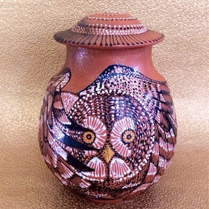 May include: A terracotta-colored ceramic urn with a detailed owl design. The owl's face and wings are intricately painted with white, black, and brown patterns. The lid is also terracotta with white dot and line details.