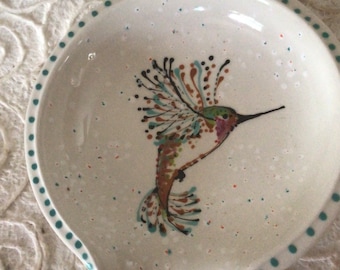 Op de afbeelding: Een witte keramische lepelsteun met een handgeschilderd kolibrie-ontwerp. De kolibrie heeft bruine, groene en blauwgroene accenten. De rand is gestippeld met blauwgroene cirkels en het oppervlak heeft gespikkelde verf.