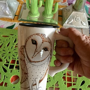 May include: White ceramic mug featuring a detailed illustration of an owl with brown and black accents. The mug has a black rim and a large, curved handle. The mug is held by a person's hand. Decorative cactus salt and pepper shakers are in the background.