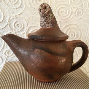 May include: A brown ceramic teapot with a lid and a handle. The lid is topped with a detailed owl figurine. The teapot has a spout and a curved handle. The background is a textured, off-white surface.