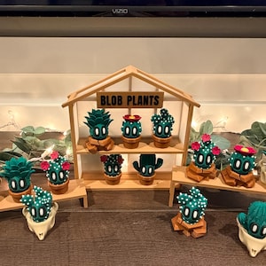 May include: A collection of whimsical "Blob Plants" cactus figurines, each with a unique design and color scheme. The display includes a wooden shelf with the text "BLOB PLANTS" and several individual display stands. The cacti are in various pots and containers.