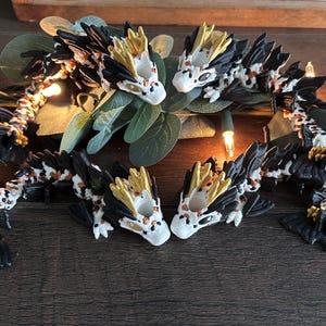 May include: A collection of handcrafted dragon figurines. The dragons feature white, black, and gold colouring with intricate details. They are arranged on a dark wooden surface with greenery and fairy lights.