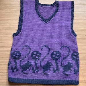 Pu&ograve; includere: Gilet di maglia viola con scollo a V e bordatura blu scuro. La parte inferiore del gilet presenta un motivo di funghi e piccoli animali blu scuro.
