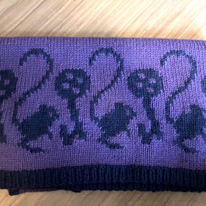 Pu&ograve; includere: Coperta in maglia viola con un motivo di gatti neri. La coperta ha un bordo blu scuro.