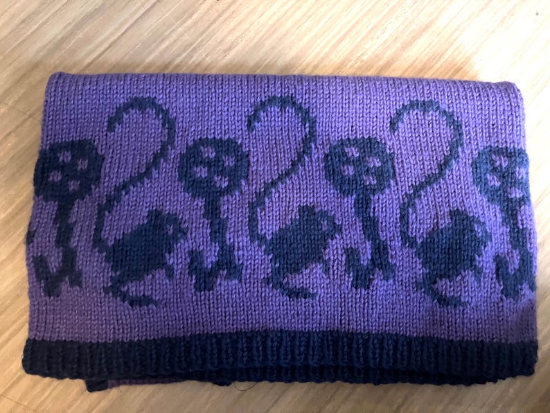 Pu&ograve; includere: Coperta in maglia viola con un motivo di gatti neri. La coperta ha un bordo blu scuro.